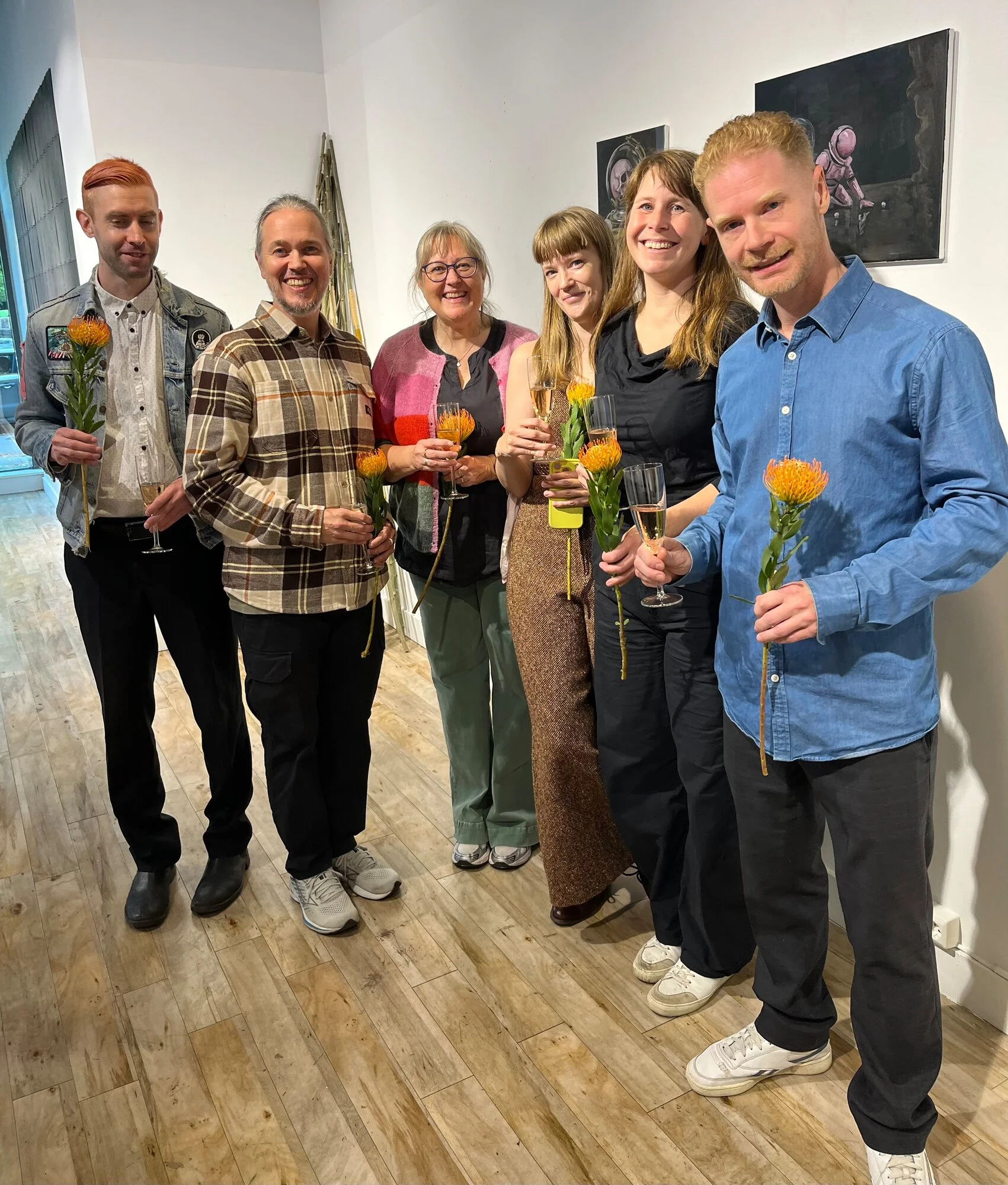 Seks personer står smilende i galleriet med hver sin oransje blomst og et glass, foran malerier på den hvite veggen.